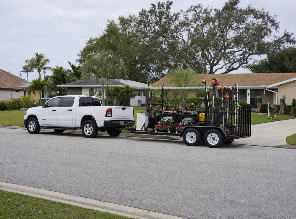 A pickup and open equipment trailer parked at the curb outside a home on a residential street.